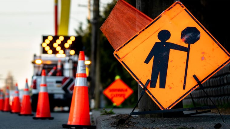 An orange road construction sign showing a worker with a stop sign stands beside traffic cones and a utility truck, indicating active road work ahead.